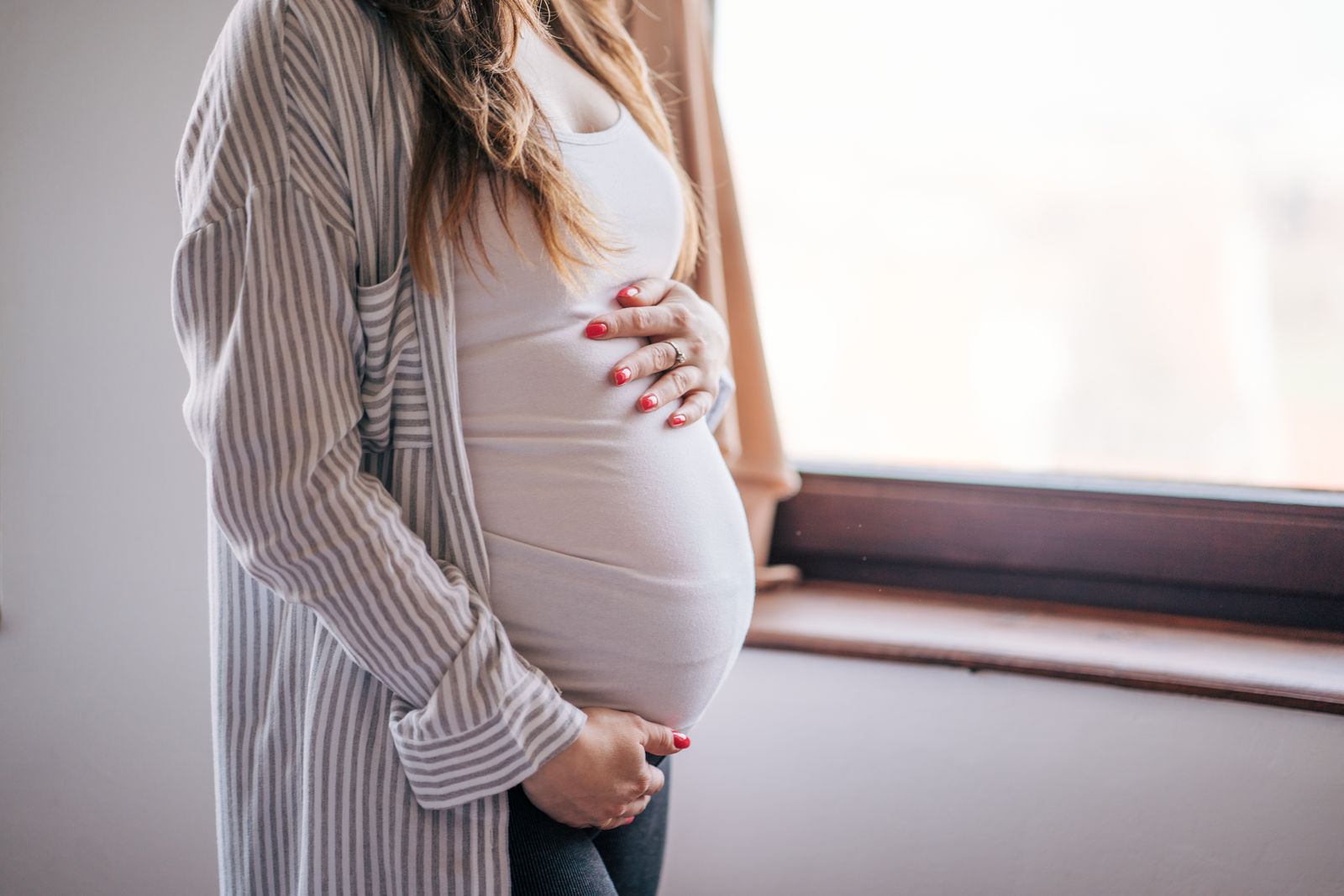 Schwangere Frau am Fenster, Hände auf dem Bauch